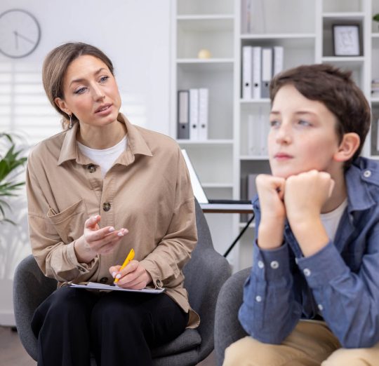 Pensive teenage guy at an appointment with a female psychologist, sitting in a modern office, immersed in his thoughts, consulting a psychotherapist, teenage problems bullying at school.